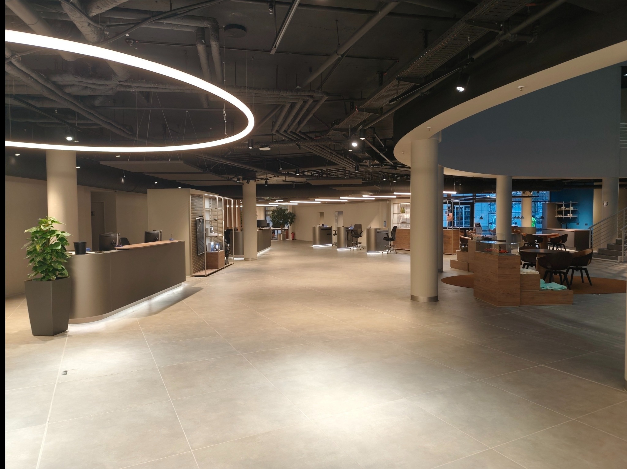 Nowoczesne wnętrze biurowe z recepcją, okrągłą lampą sufitową i widocznymi instalacjami pod sufitem. Minimalistyczny design, jasna podłoga, kolumny i rośliny doniczkowe.