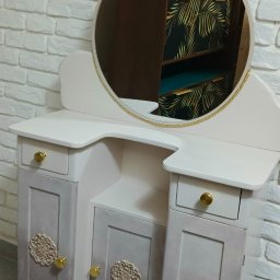 Odnowiona toaletka w stylu vintage z okrągłym lustrem i dekoracyjnymi aplikacjami na frontach szafek, na tle ściany z białej cegły.