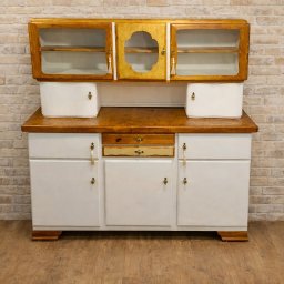 Odrestaurowany kredens w stylu vintage z przeszklonymi szafkami i białymi frontami, na tle ceglanej ściany i drewnianej podłogi. Widoczne detale wykończenia i ozdobne uchwyty.