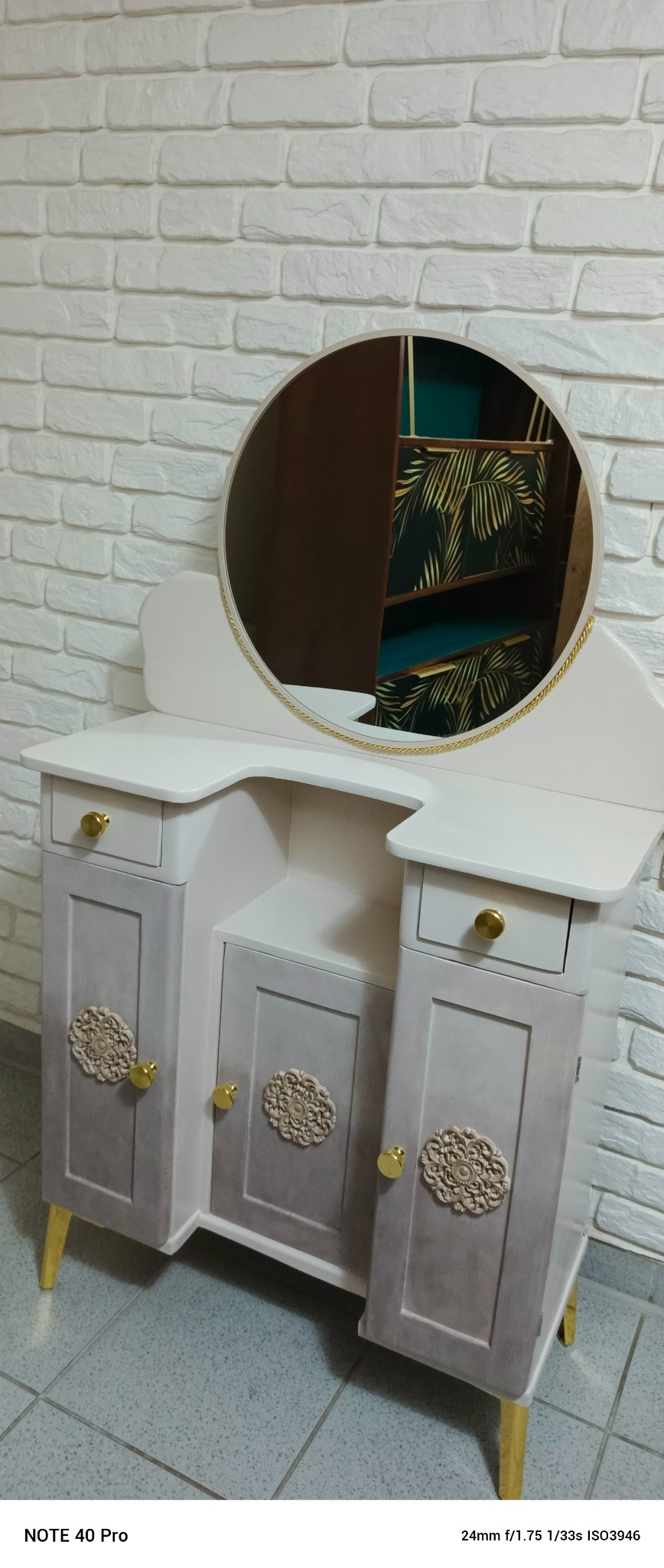 Odnowiona toaletka w stylu vintage z okrągłym lustrem i dekoracyjnymi aplikacjami na frontach szafek, na tle ściany z białej cegły.