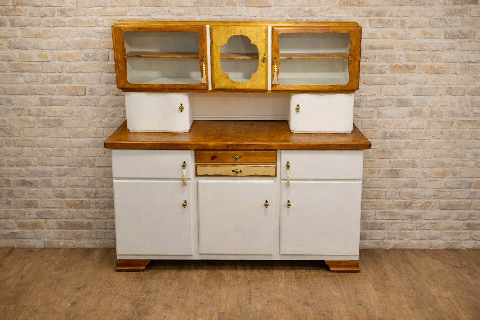 Odrestaurowany kredens w stylu vintage z przeszklonymi szafkami i białymi frontami, na tle ceglanej ściany i drewnianej podłogi. Widoczne detale wykończenia i ozdobne uchwyty.