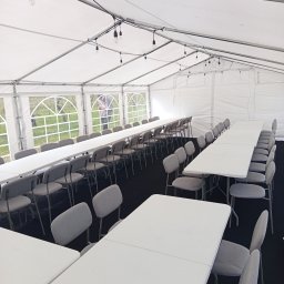 JM EVENT - Wnętrze białego namiotu eventowego z rzędami białych stołów i szarych krzeseł, oświetlonego girlandami żarówek. Za oknami widoczna zieleń.