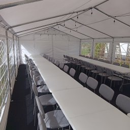 JM EVENT - Wnętrze białego namiotu bankietowego z długimi stołami i szarymi krzesłami, oświetlonego girlandami żarówek. Widok z perspektywy, czarna podłoga.
