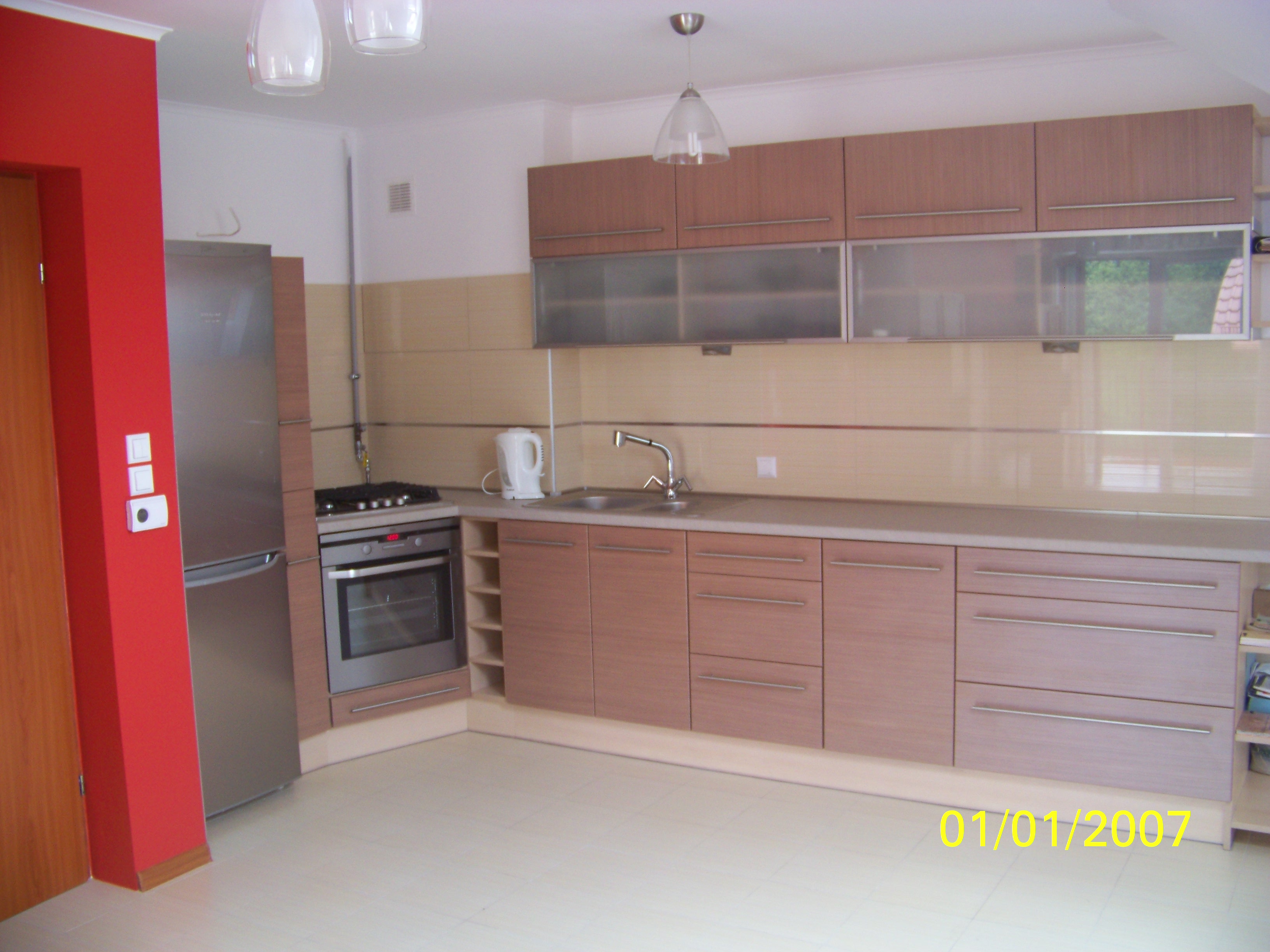 Jasna kuchnia z drewnopodobnymi frontami, stalowymi uchwytami, i beżowymi płytkami nad blatem, wyposażona w sprzęt AGD ze stali nierdzewnej: lodówkę, piekarnik i płytę gazową.