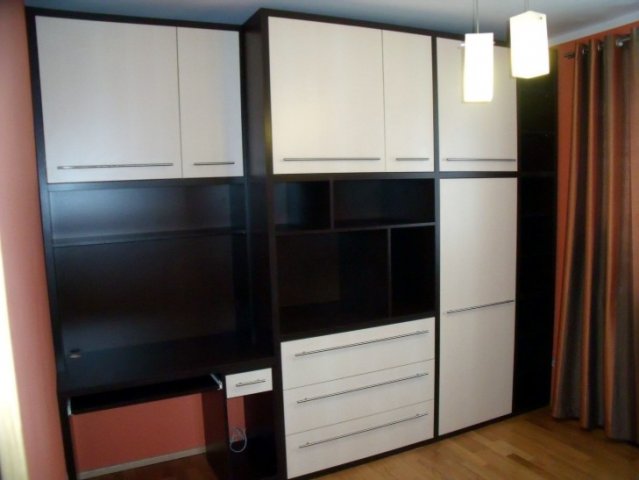 Zabudowa ścienna z biurkiem, szafkami i szufladami w kolorach bieli i ciemnego brązu, oświetlona wiszącymi lampami.