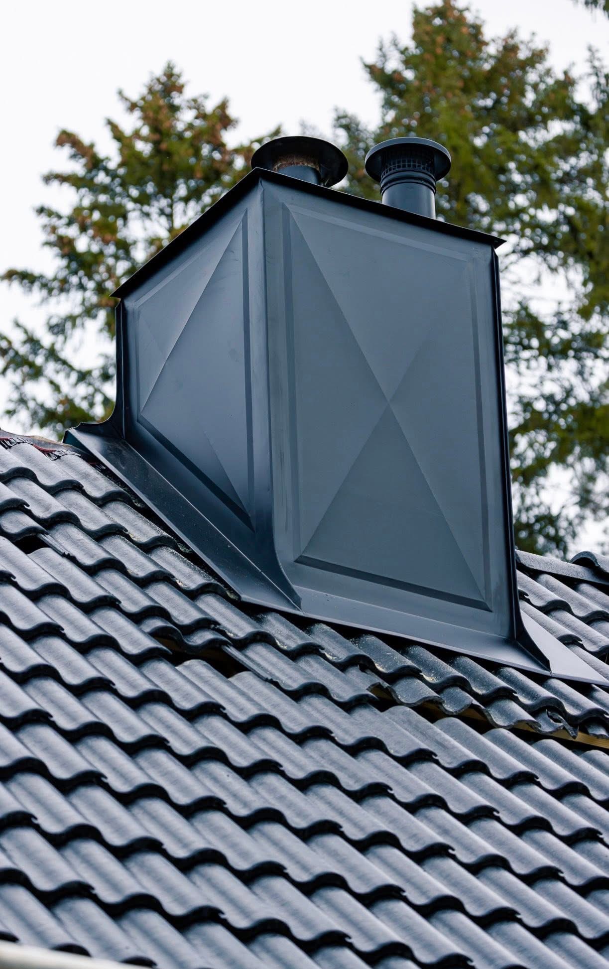 Szczegółowe ujęcie czarnego, metalowego komina z dwoma przewodami wentylacyjnymi na dachu z ciemnej dachówki ceramicznej, na tle zielonego drzewa.