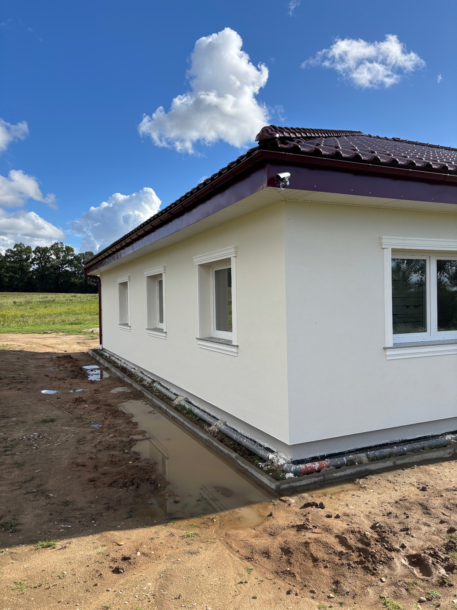 Elewacja domu jednorodzinnego z brązowym dachem i białymi oknami, widok z zewnątrz w słoneczny dzień. Wokół domu ziemia i kałuże.