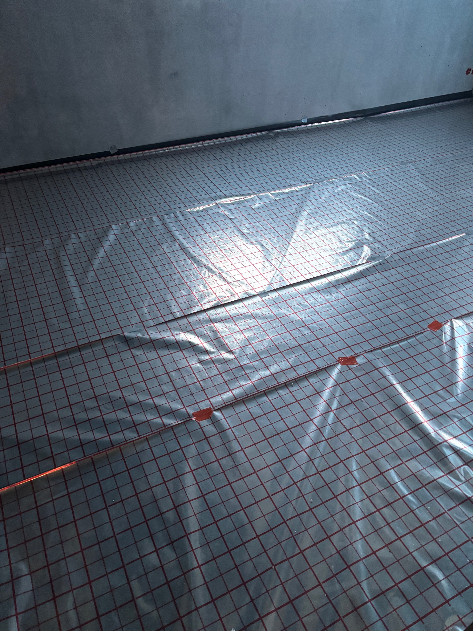 Instalacja ogrzewania podłogowego: widok na ułożone kable grzewcze na folii z siatką, przymocowane pomarańczową taśmą, gotowe do zalania wylewką.