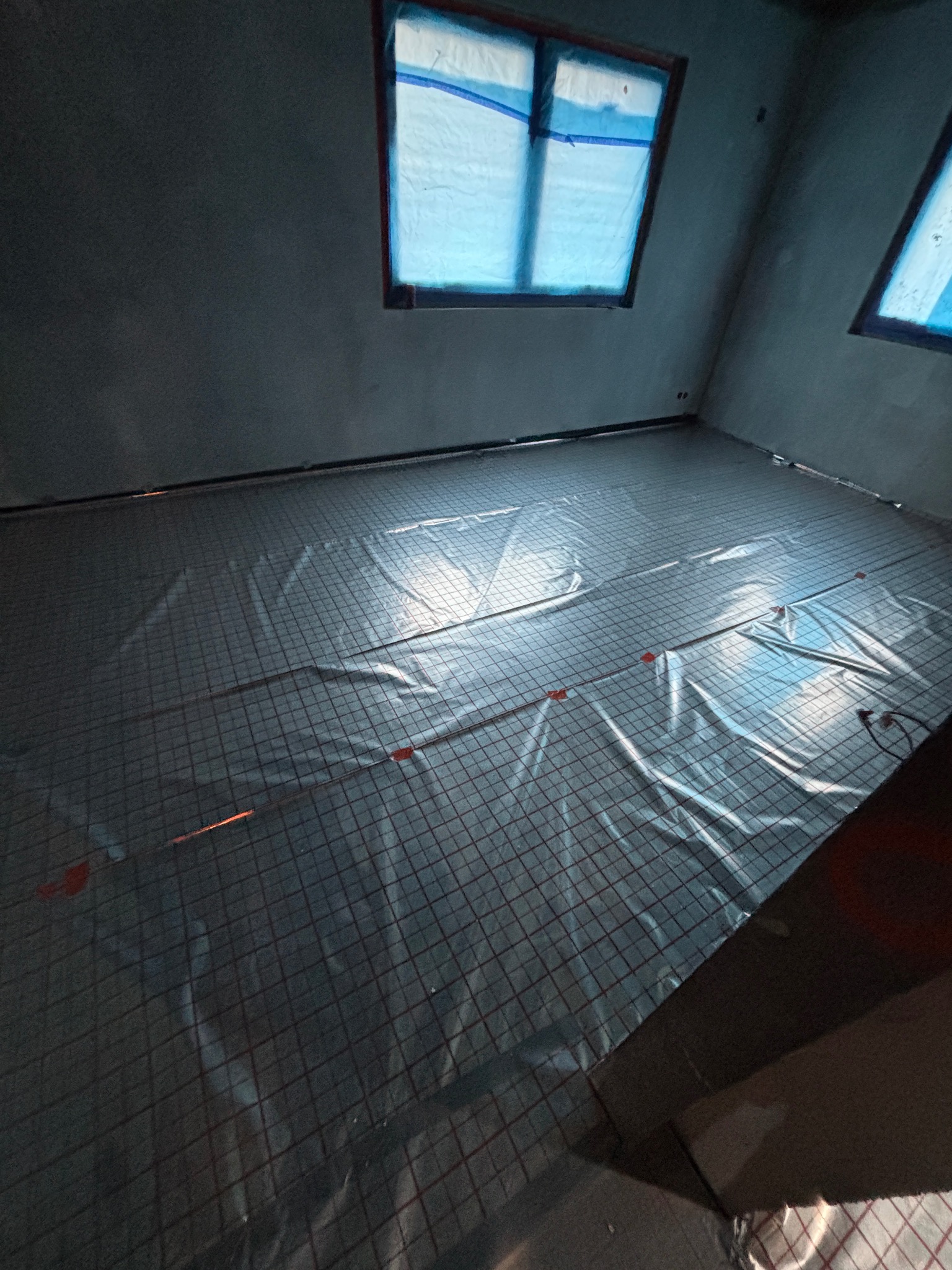 Przygotowanie do wylewki: rozłożona siatka z ogrzewaniem podłogowym w surowym pomieszczeniu z oknami zabezpieczonymi folią. Widok z góry, ciemne ściany.