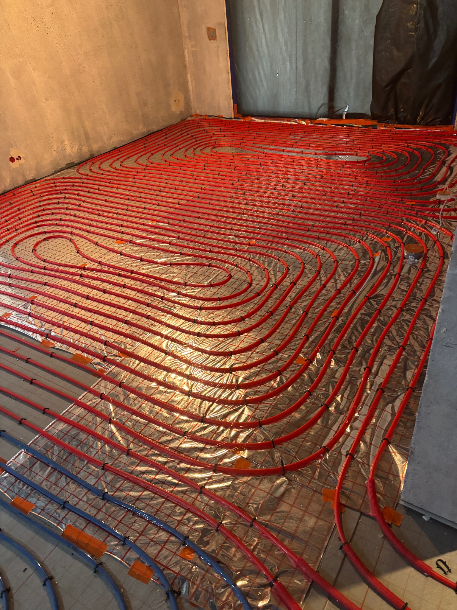 Czerwone i niebieskie rury ogrzewania podłogowego ułożone na folii izolacyjnej w surowym wnętrzu. Widoczne mocowania rur do siatki. Etap instalacji przed wylewką.