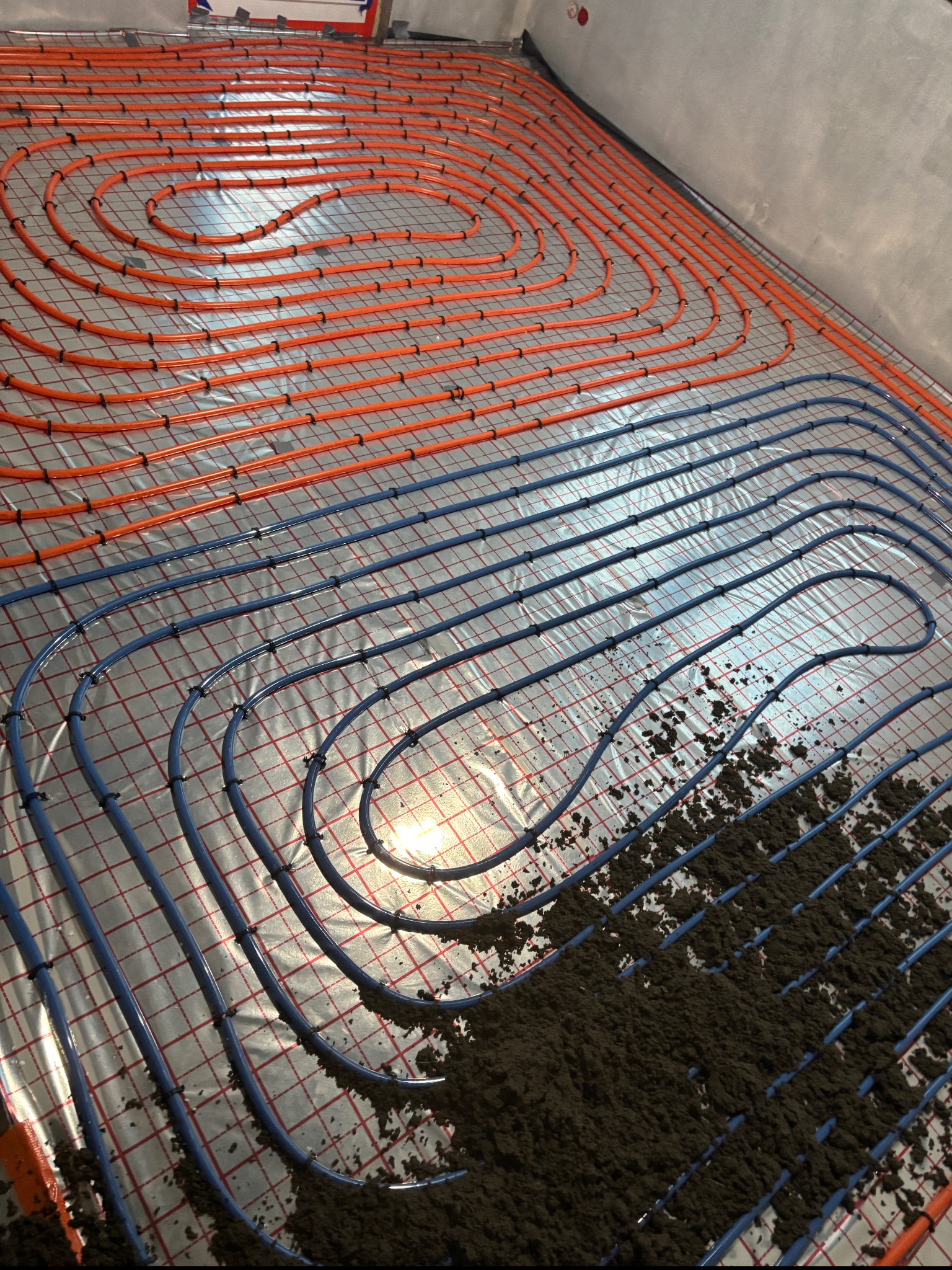 Rozłożona instalacja ogrzewania podłogowego z rurami w kolorze pomarańczowym i niebieskim na folii izolacyjnej z widocznymi fragmentami zaprawy w dolnej części kadru.