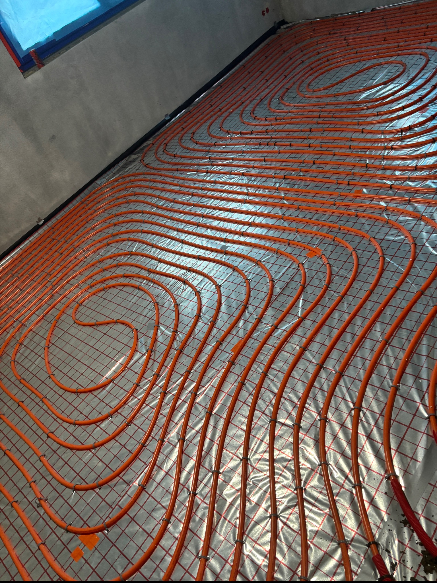 Pomarańczowe rury ogrzewania podłogowego ułożone spiralnie na srebrnej folii z siatką, widoczne fragmenty ściany i okna. Instalacja przed wylaniem posadzki.