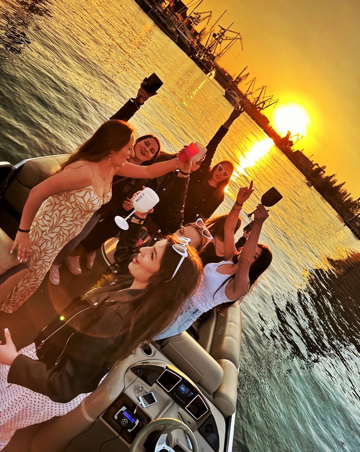 Grupa kobiet świętuje na łodzi podczas zachodu słońca, trzymając kieliszki i wznosząc toasty. W tle widoczne dźwigi portowe i złocista woda.