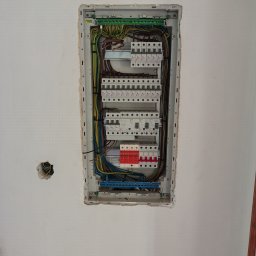 EL-KAR Karol Napora - Rozdzielnia elektryczna w ścianie, widoczne kable i zabezpieczenia. Ściana w trakcie wykończenia, widoczne otwory na gniazdka. Ujęcie z bliska, oświetlenie naturalne.