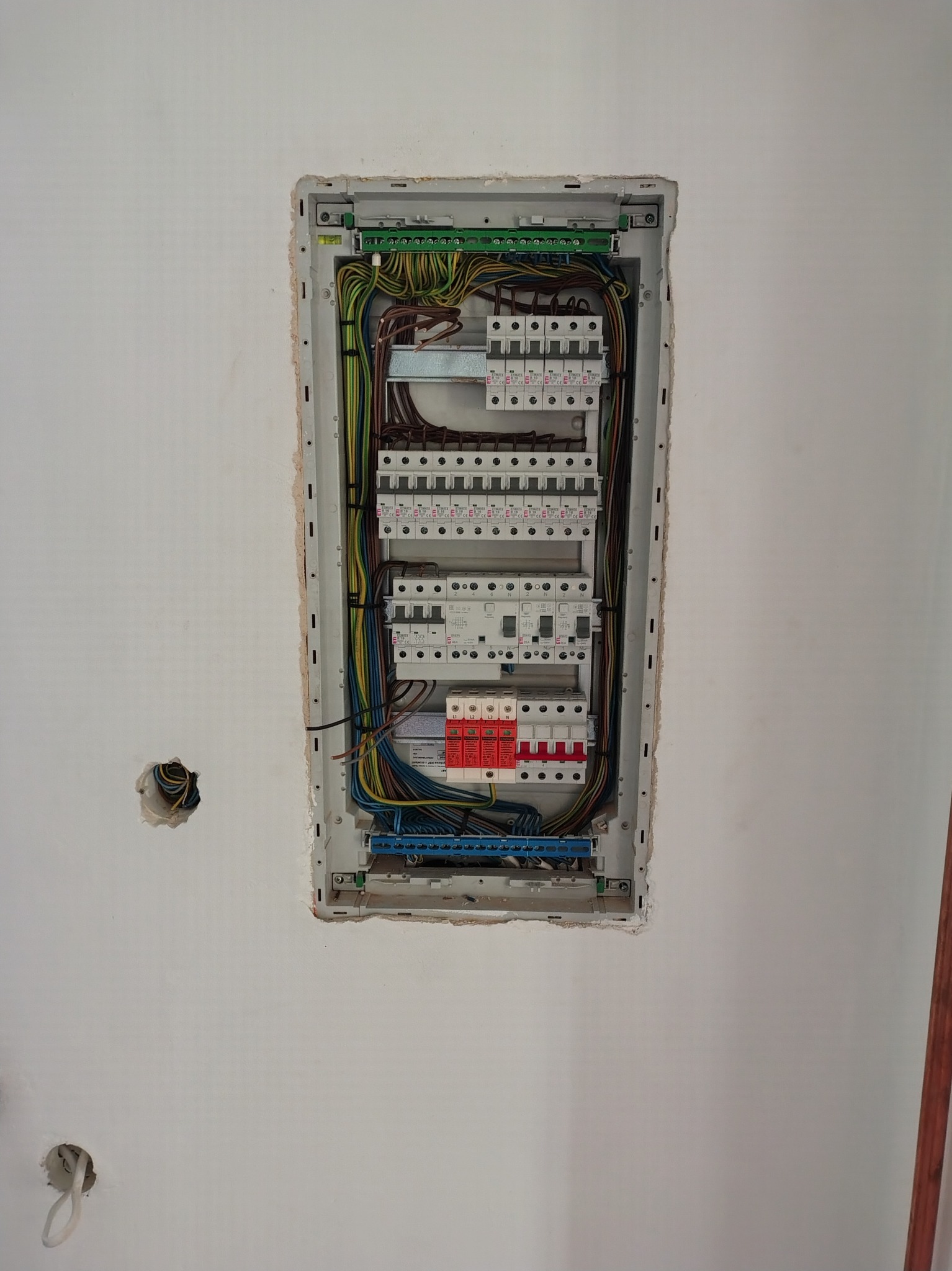 Rozdzielnia elektryczna w ścianie, widoczne kable i zabezpieczenia. Ściana w trakcie wykończenia, widoczne otwory na gniazdka. Ujęcie z bliska, oświetlenie naturalne.
