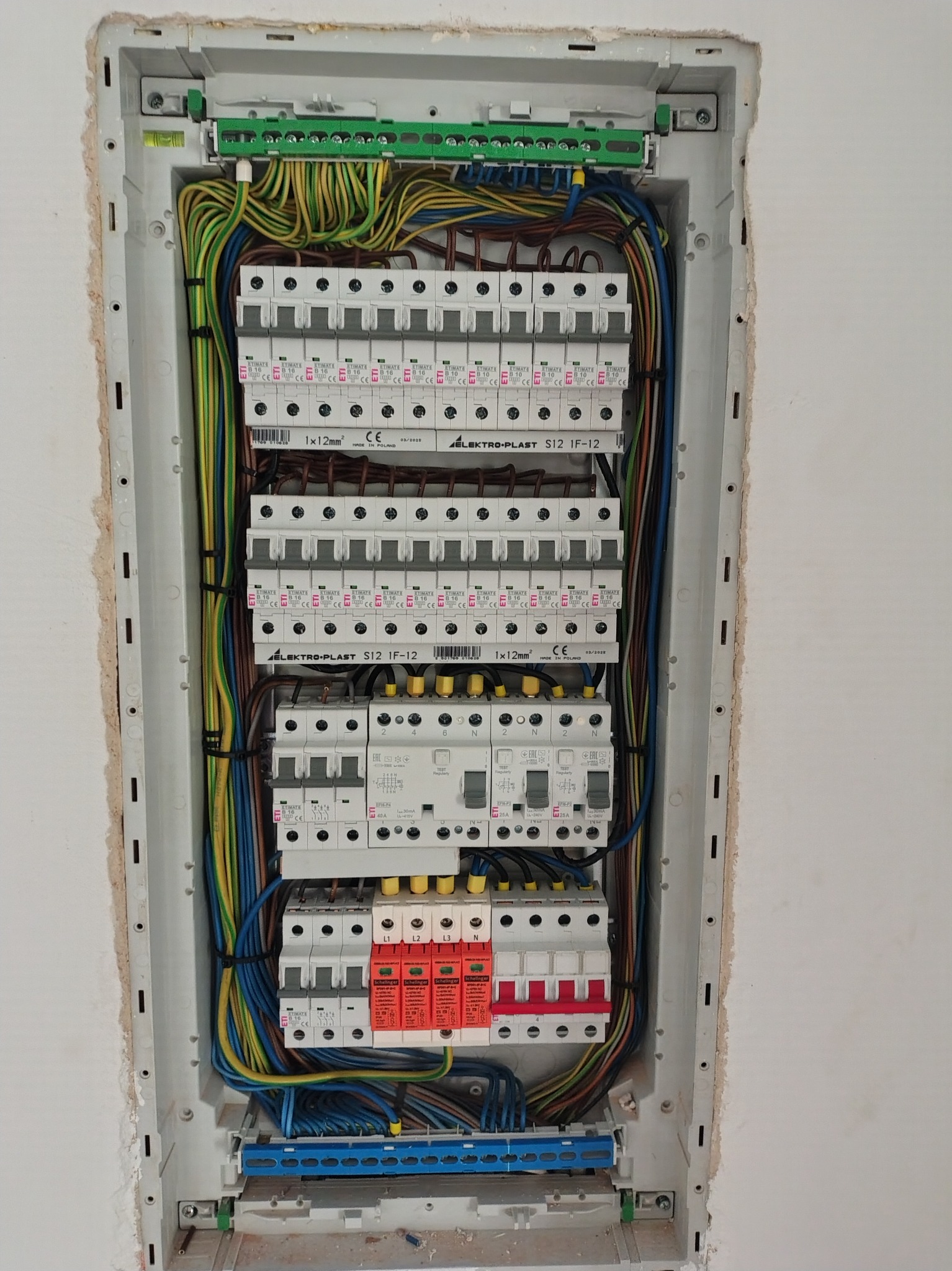 Rozdzielnica elektryczna w obudowie, widoczne zabezpieczenia, przewody w różnych kolorach, listwy zaciskowe. Instalacja w trakcie montażu, ściana z widocznymi śladami prac.