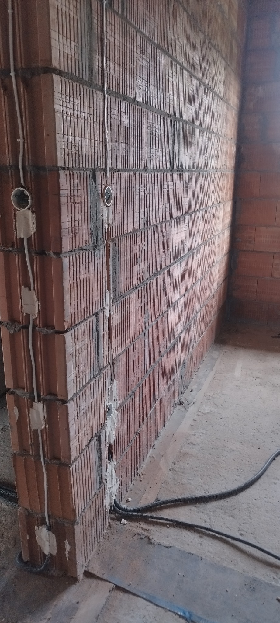 Czerwona ściana z cegły dziurawki z widocznymi przewodami elektrycznymi i puszkami instalacyjnymi. Przewody prowadzone wzdłuż ściany, częściowo zamaskowane gipsem.