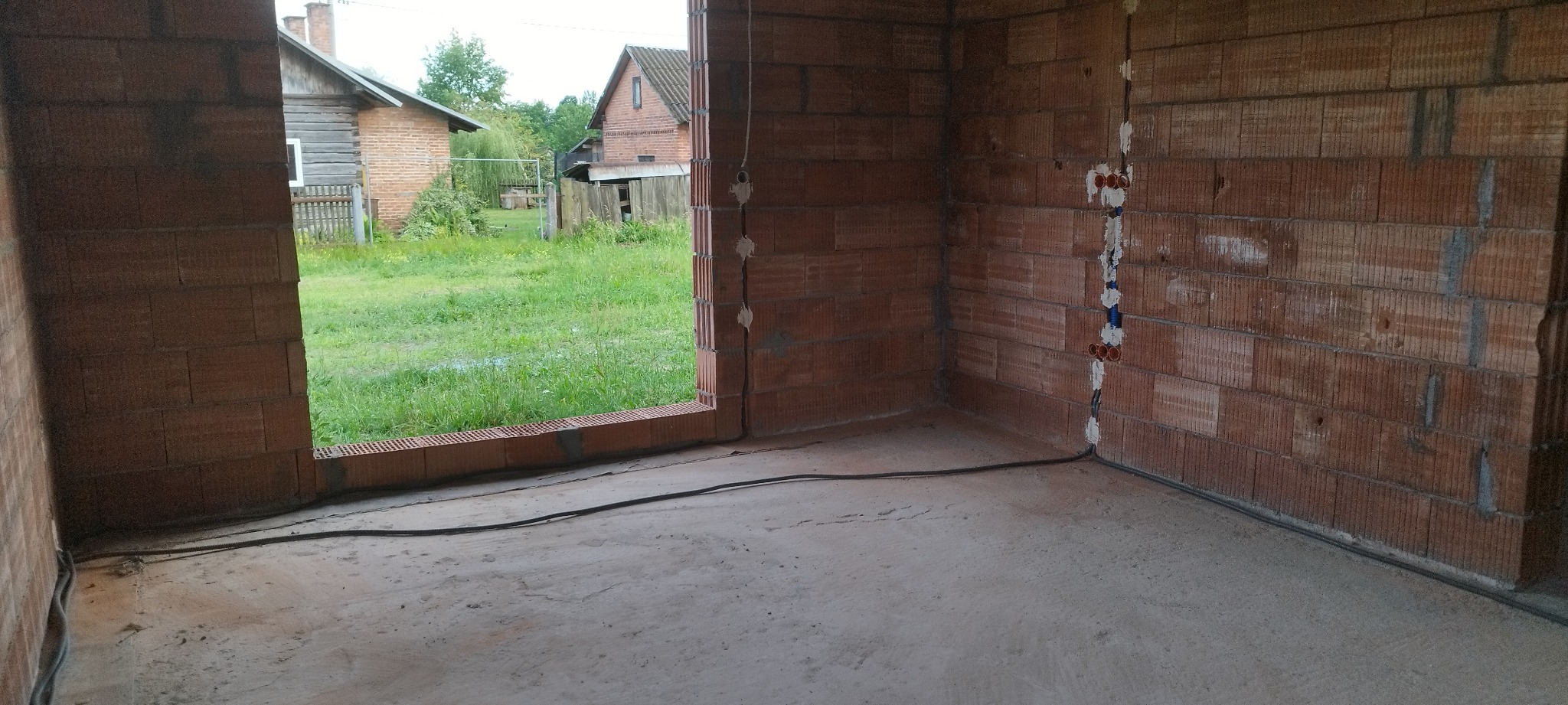 Surowy stan wnętrza z instalacją elektryczną w bruzdach ścian z czerwonej cegły. Widoczne kable i puszki. W tle zieleń i zabudowania przez otwór okienny.