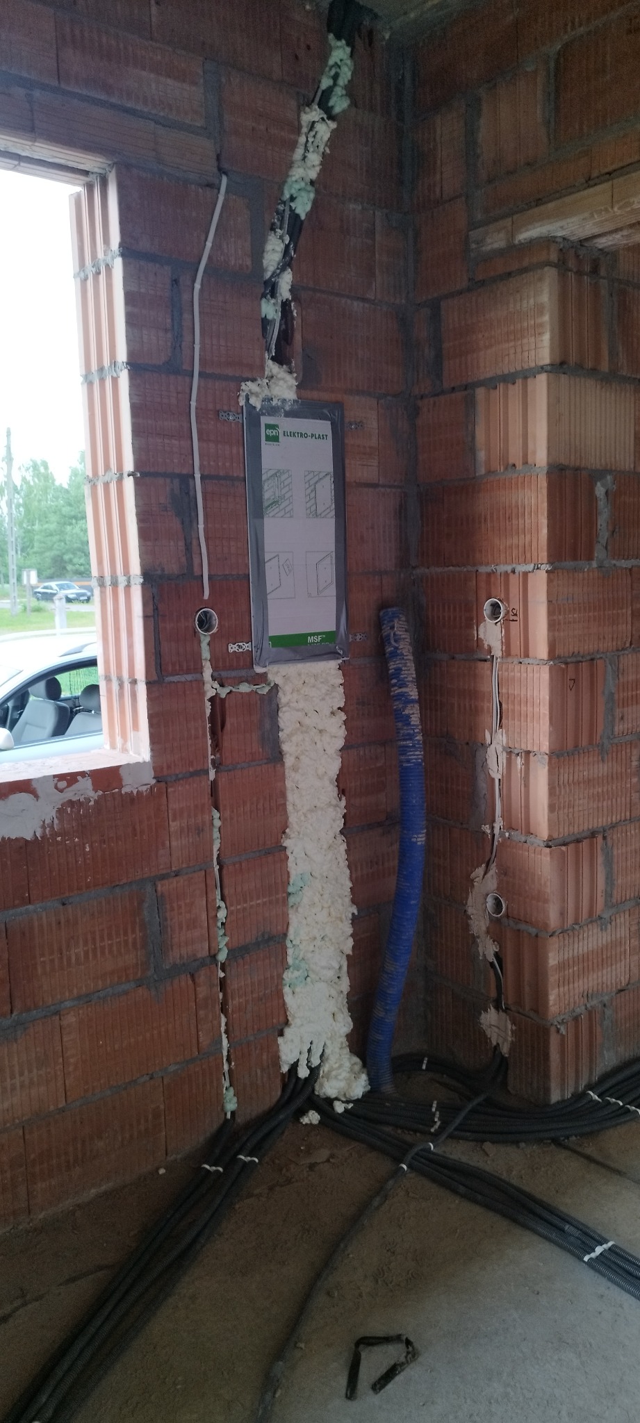 Wykute bruzdy w ceglanym murze wypełnione pianką montażową, w których umieszczono przewody. Widoczna puszka instalacyjna i peszel. Etap prac instalacyjnych w surowym budynku.
