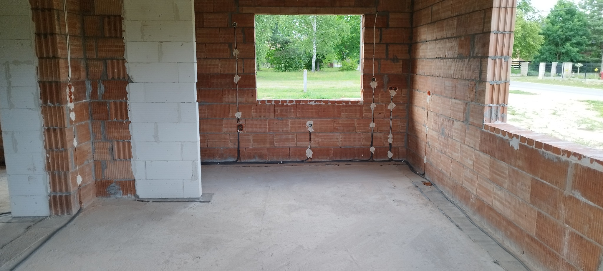 Surowy stan wnętrza z ceglanymi ścianami i widoczną instalacją elektryczną w peszlach, przygotowaną do tynkowania. Widok na zewnątrz przez okno.