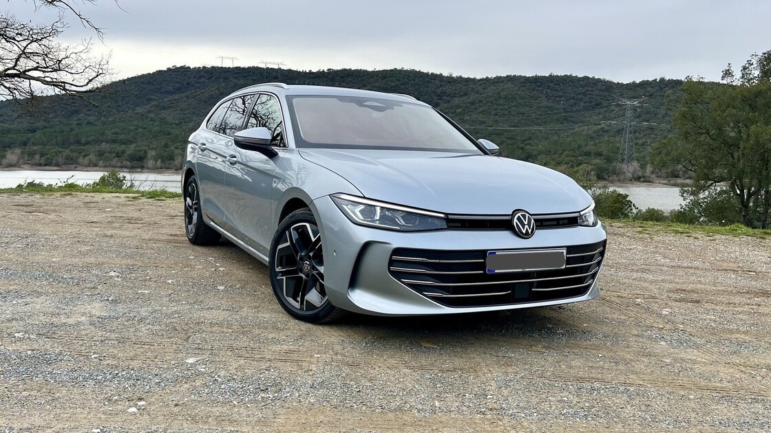 Srebrny Volkswagen kombi na żwirowym podłożu z widokiem na jezioro i zalesione wzgórza w tle. Auto z czarnymi felgami, nowoczesny design.