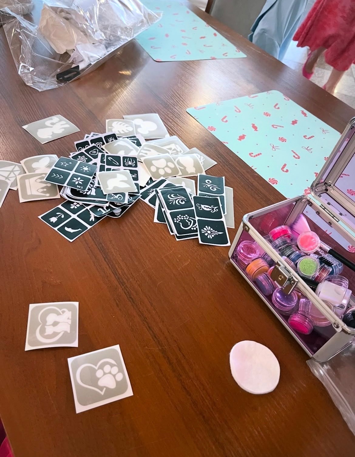 Akcesoria do malowania twarzy dla dzieci: szablony z motywami zwierząt, brokatowe farby w kuferku i gąbka na drewnianym stole z podkładkami w cukierki.