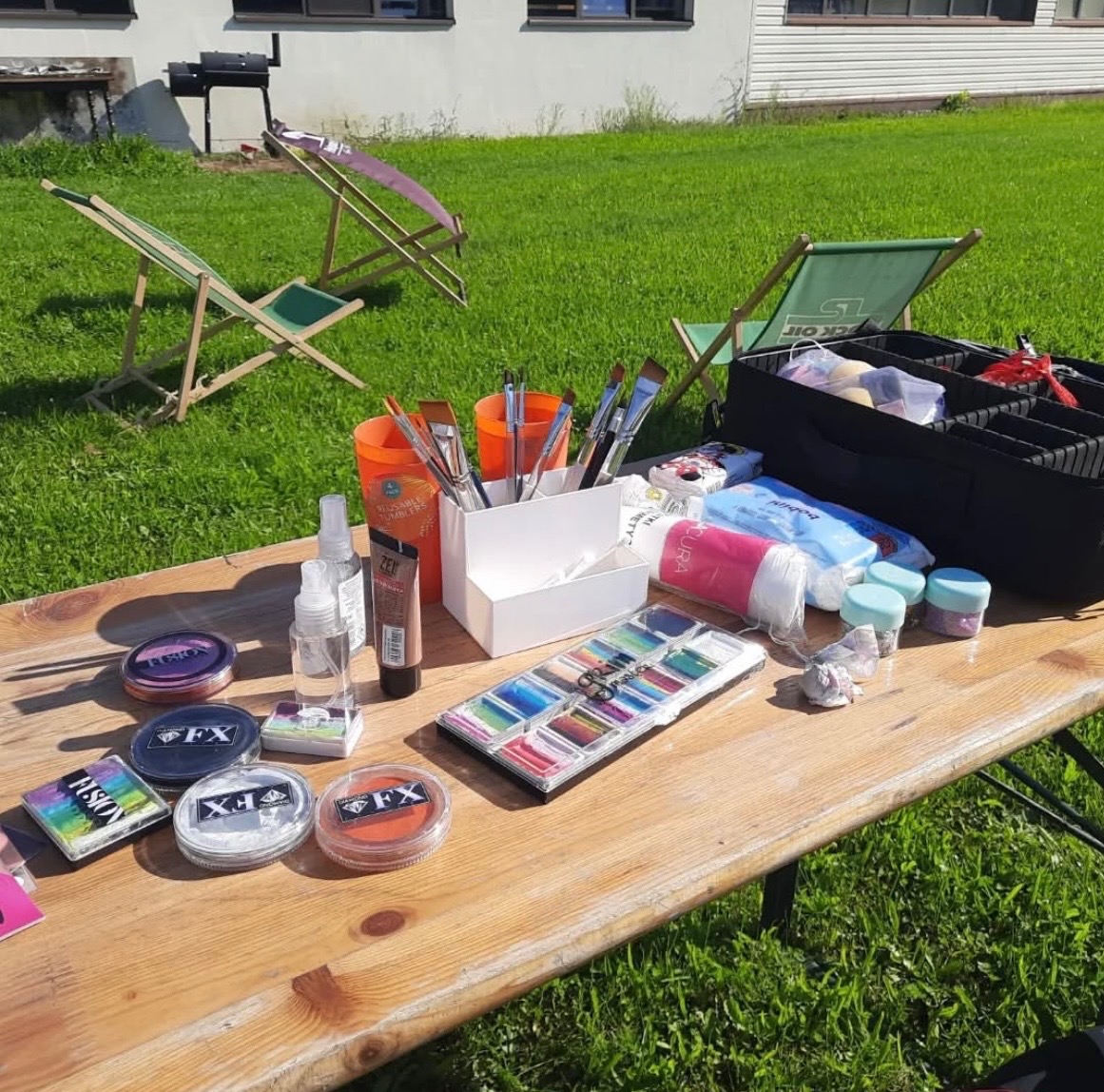 Stół z akcesoriami do malowania twarzy: farby, pędzle, kosmetyki. W tle trawa i leżaki, sugerujące plenerowe stanowisko pracy animatora.
