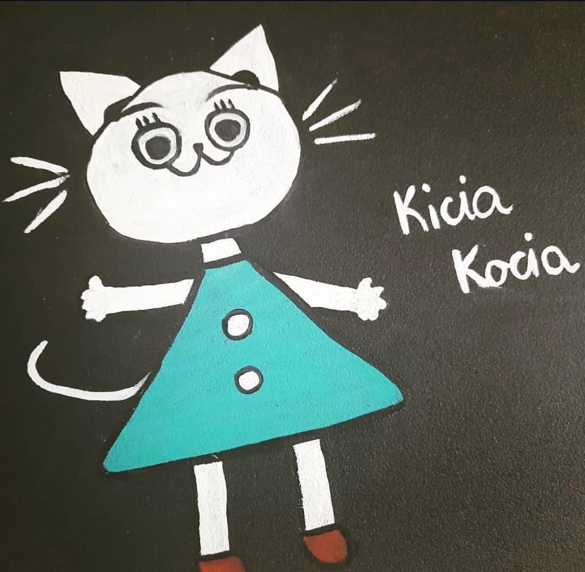 Ręcznie malowana postać Kici Koci na czarnym tle, w turkusowej sukience z białymi guzikami. Biała głowa kota z wąsami i imię postaci obok.