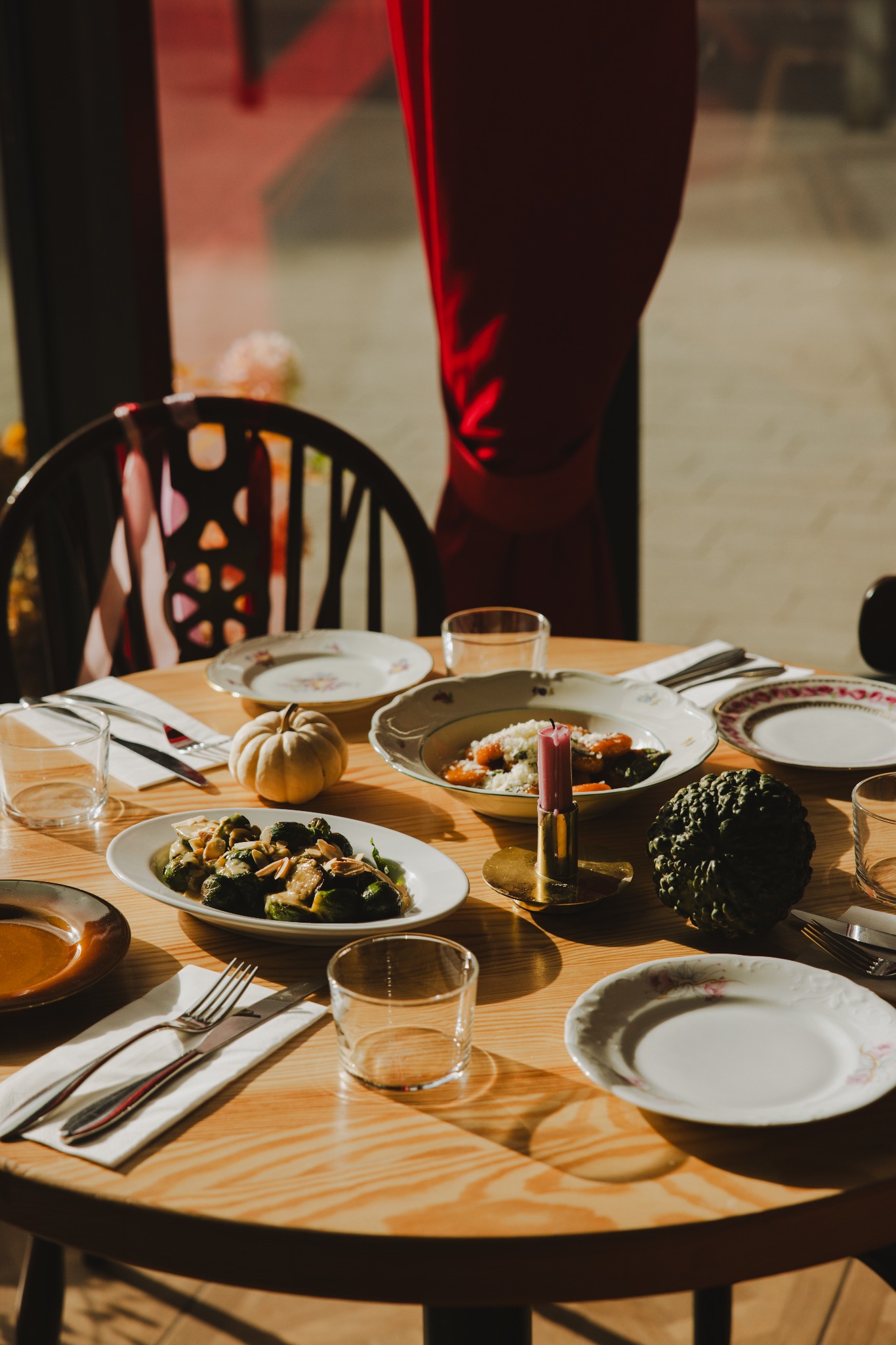 Stół z jesienną dekoracją i daniami: brukselka z migdałami, dyniowe gnocchi ze świecą. Słoneczne światło podkreśla ciepłą atmosferę spotkania.