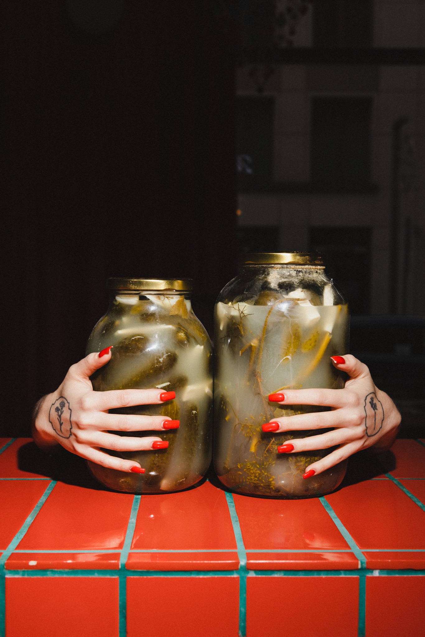 Dwie ręce z czerwonymi paznokciami obejmują słoiki z ogórkami kiszonymi na czerwonej, kafelkowej powierzchni. Widoczne tatuaże na dłoniach, ciemne tło.