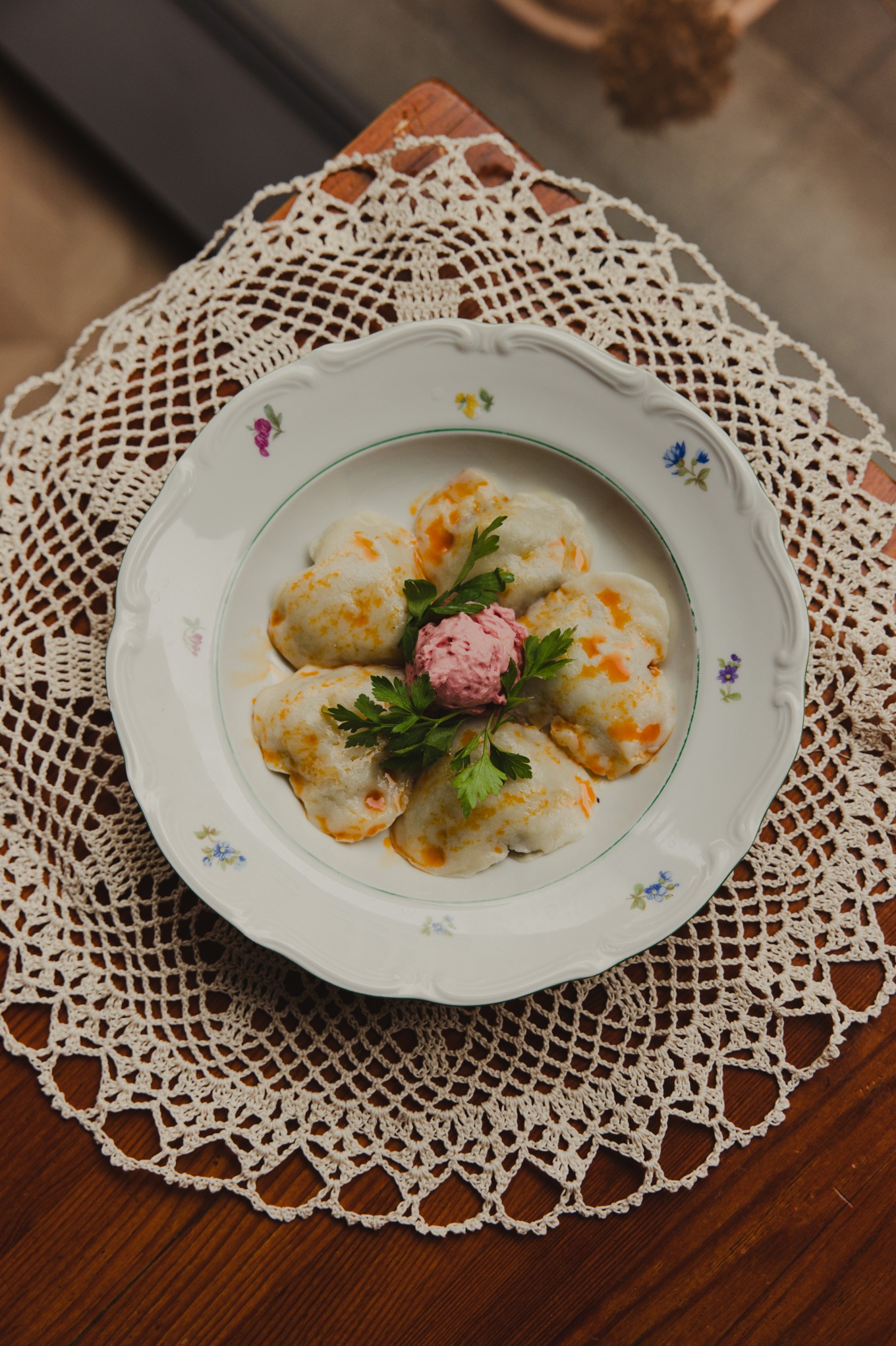 Elegancko podane pierogi z sosem i dekoracją z zieleniny na talerzu z kwiatowym motywem, umieszczonym na ażurowej serwetce na drewnianym stole.