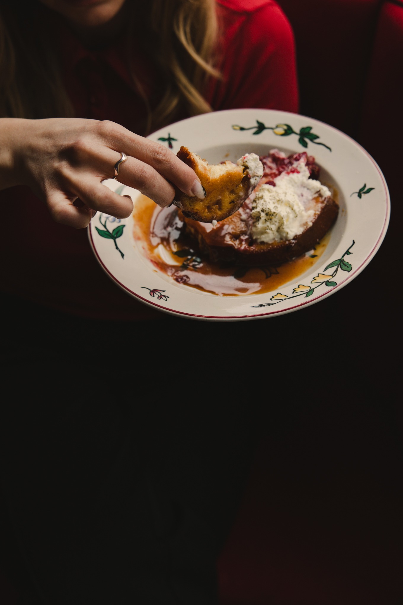 Kobieta z pierścionkiem je francuskie tosty z bitą śmietaną i sosem na talerzu z motywem kwiatowym. Czerwona bluzka i ciemne tło.