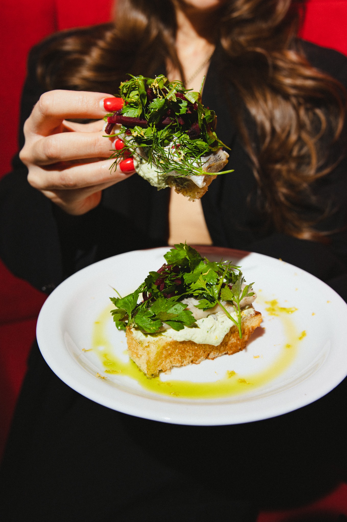 Kanapki z rybą i ziołami na białym talerzu, trzymane w dłoni z czerwonymi paznokciami, na czerwonym tle. Elegancki catering.