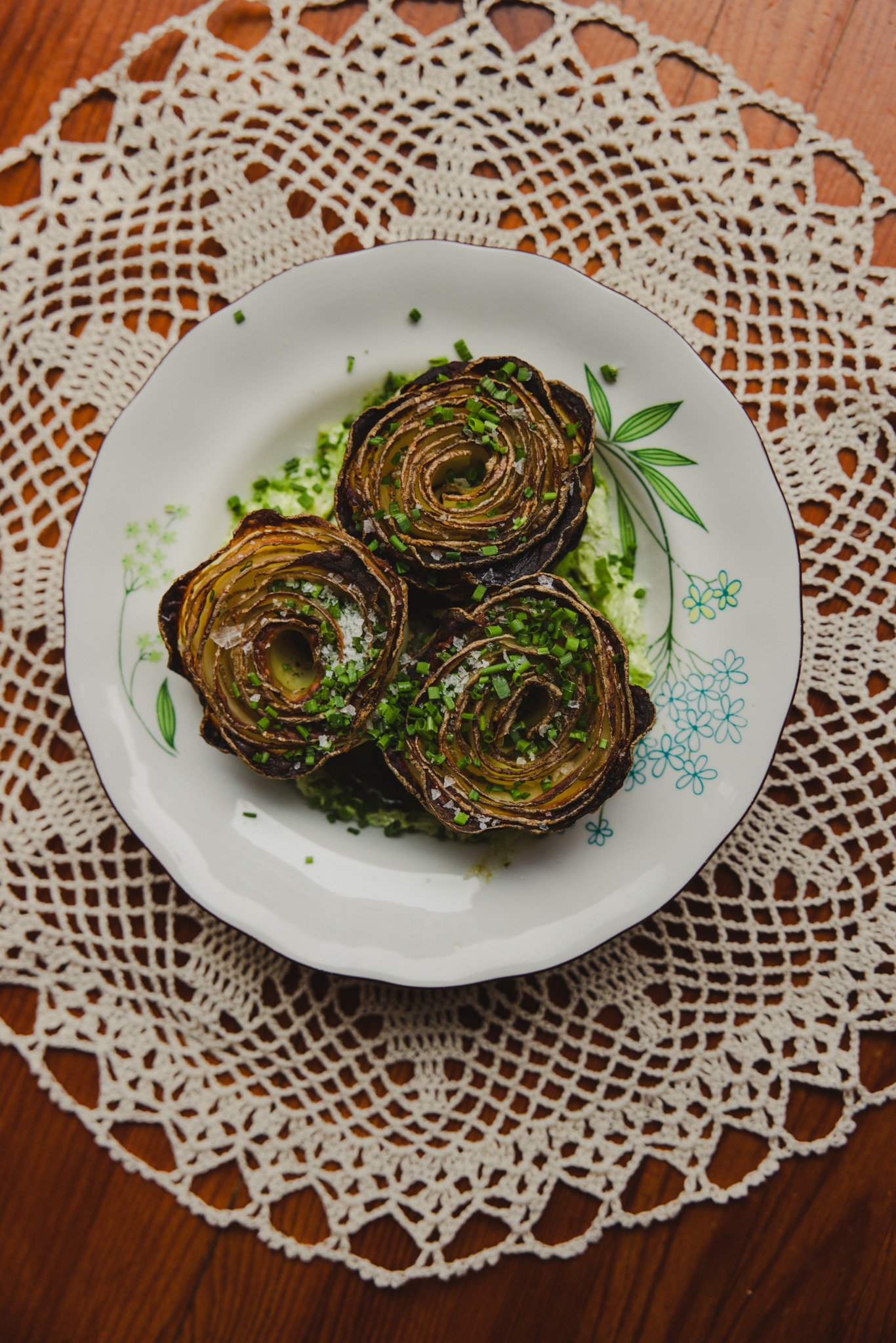 Trzy pieczone karczochy ułożone na talerzu z zielonym sosem i posypane szczypiorkiem, na drewnianym stole z serwetą. Karczochy mają spiralny kształt.