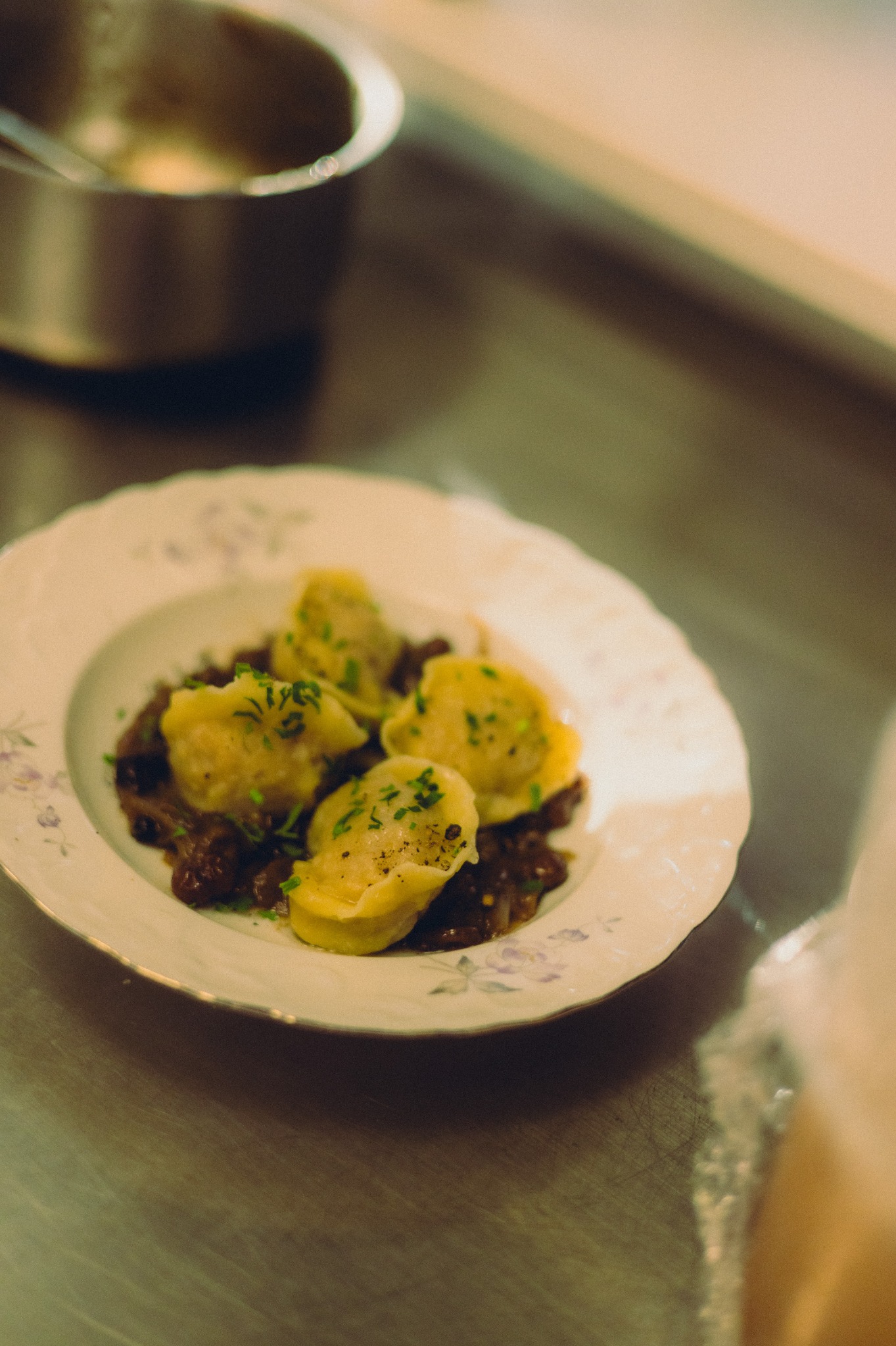 Delikatne pierożki tortellini, skropione świeżym koperkiem, ułożone na ciemnym sosie na porcelanowym talerzu z kwiatowym motywem. W tle metalowy garnek.