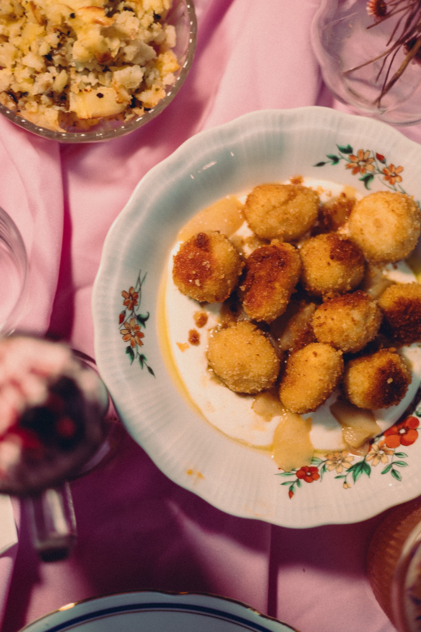 Kluski śląskie na talerzu z kwiatowym wzorem, otoczone różowym obrusem i innymi naczyniami. Ujęcie z góry, ciepłe światło, domowa atmosfera.