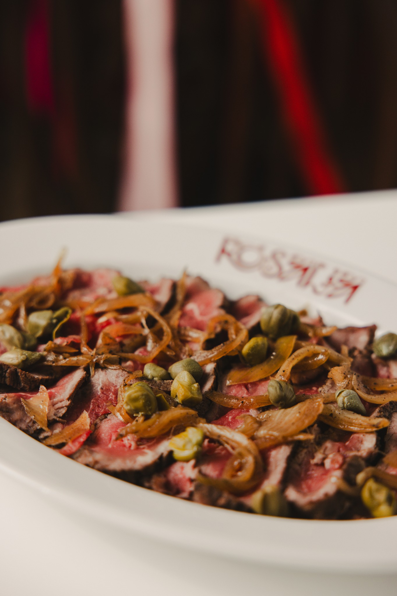 Carpaccio wołowe z kaparami i karmelizowaną cebulą na białym talerzu z logo restauracji, w ciepłym oświetleniu.
