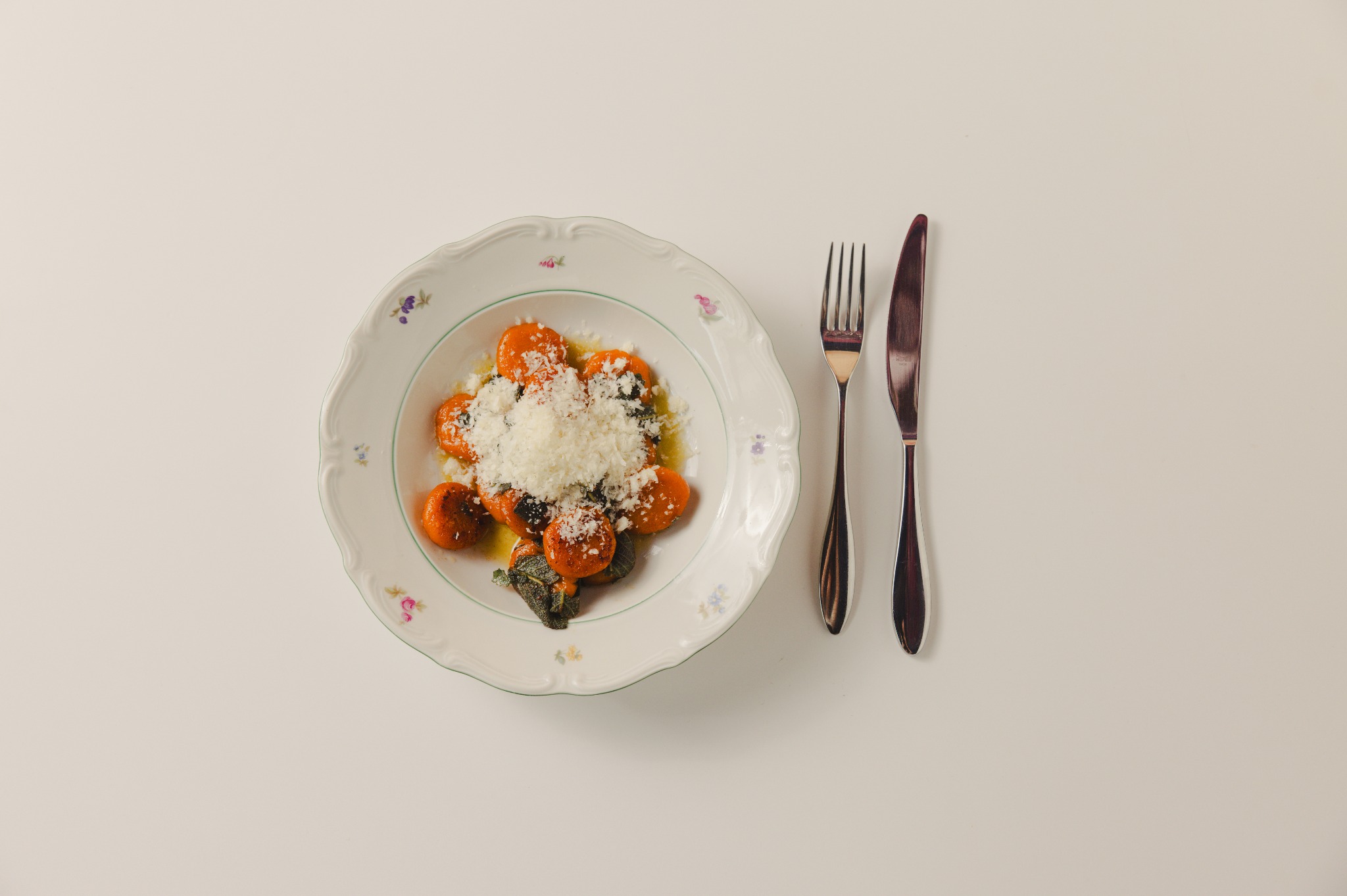 Pomarańczowe gnocchi z szałwią i parmezanem na talerzu z kwiatowym wzorem, obok widelec i nóż na białym tle. Ujęcie z góry, miękkie światło.