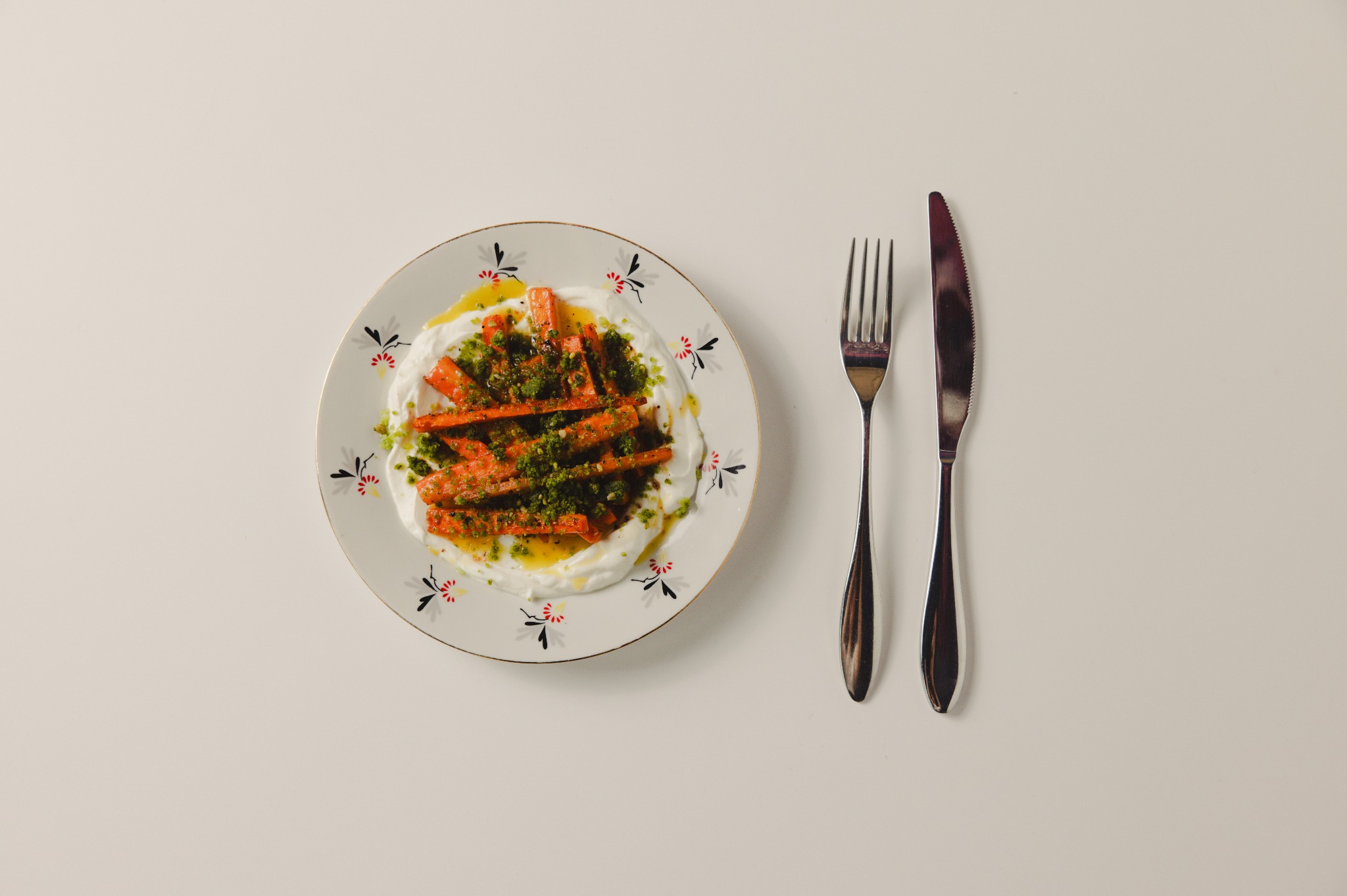 Apetyczna kompozycja: pieczona marchewka z pistacjami na kremowym jogurcie, podana na talerzu z kwiatowym wzorem, obok widelec i nóż na białym tle.