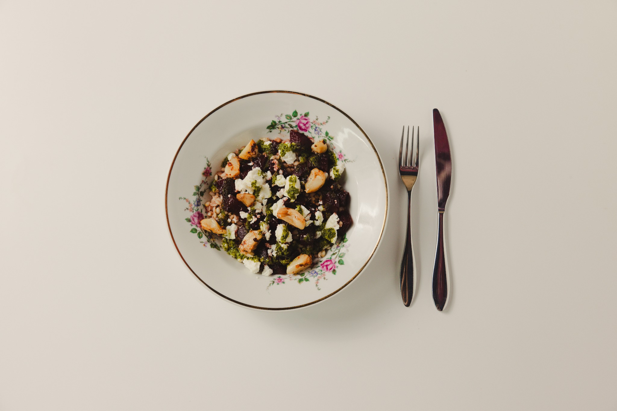Sałatka z buraków, sera feta i pesto na talerzu z kwiatowym wzorem, obok widelec i nóż na białym tle. Minimalistyczne ujęcie z góry, stonowana kolorystyka.