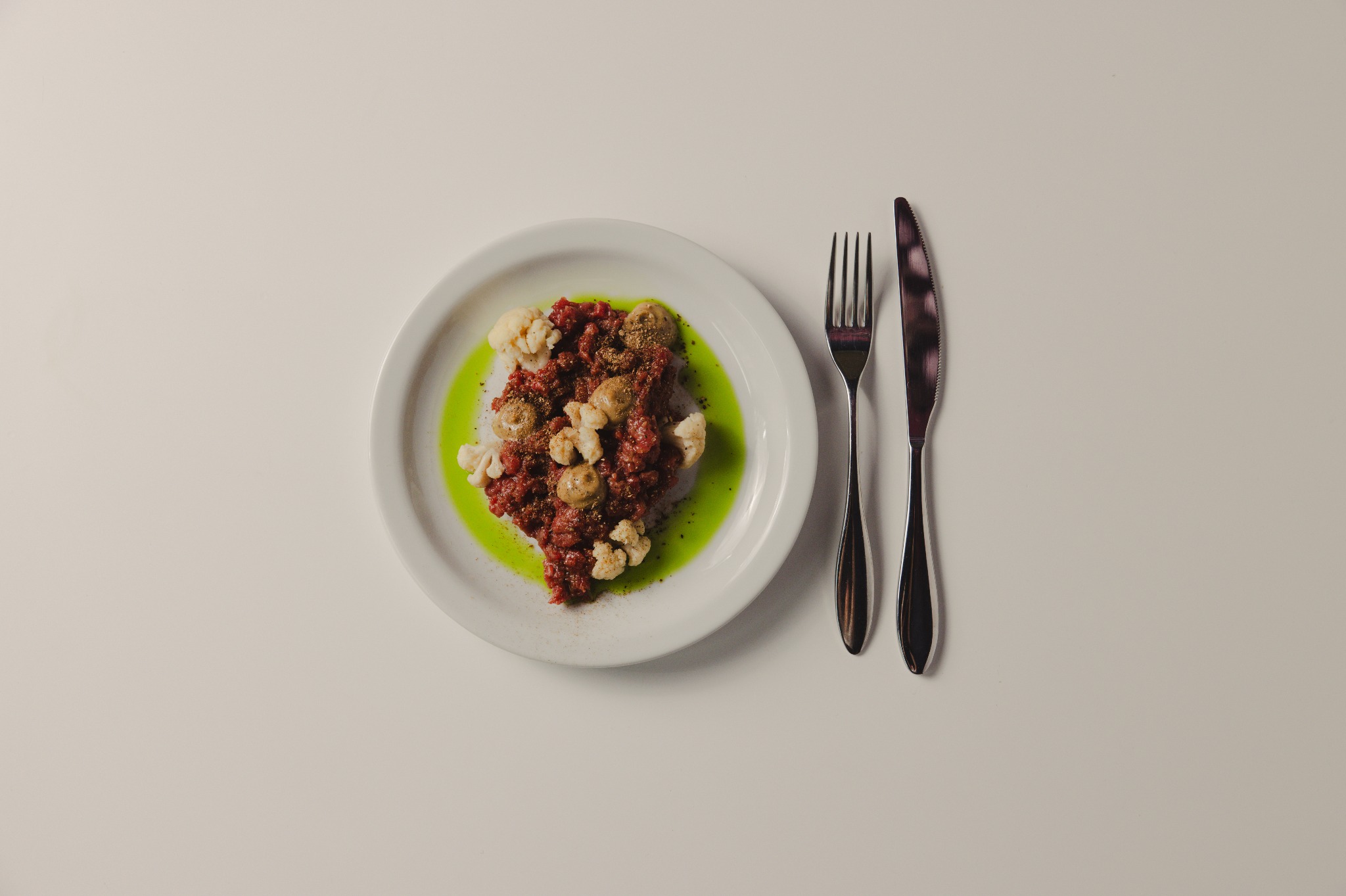 Wykwintny tatar wołowy z kalafiorem i sosem na białym talerzu, obok widelec i nóż. Elegancka kompozycja kulinarna na jasnym tle, idealna na catering.