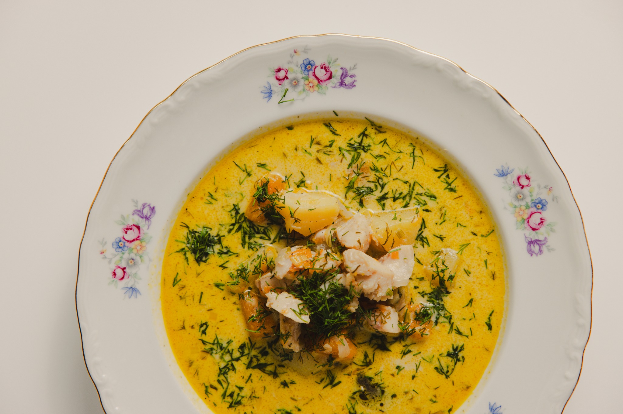 Zupa rybna z koperkiem i ziemniakami w białym talerzu z kwiatowym wzorem i złotym obramowaniem, widok z góry, na jasnym tle.