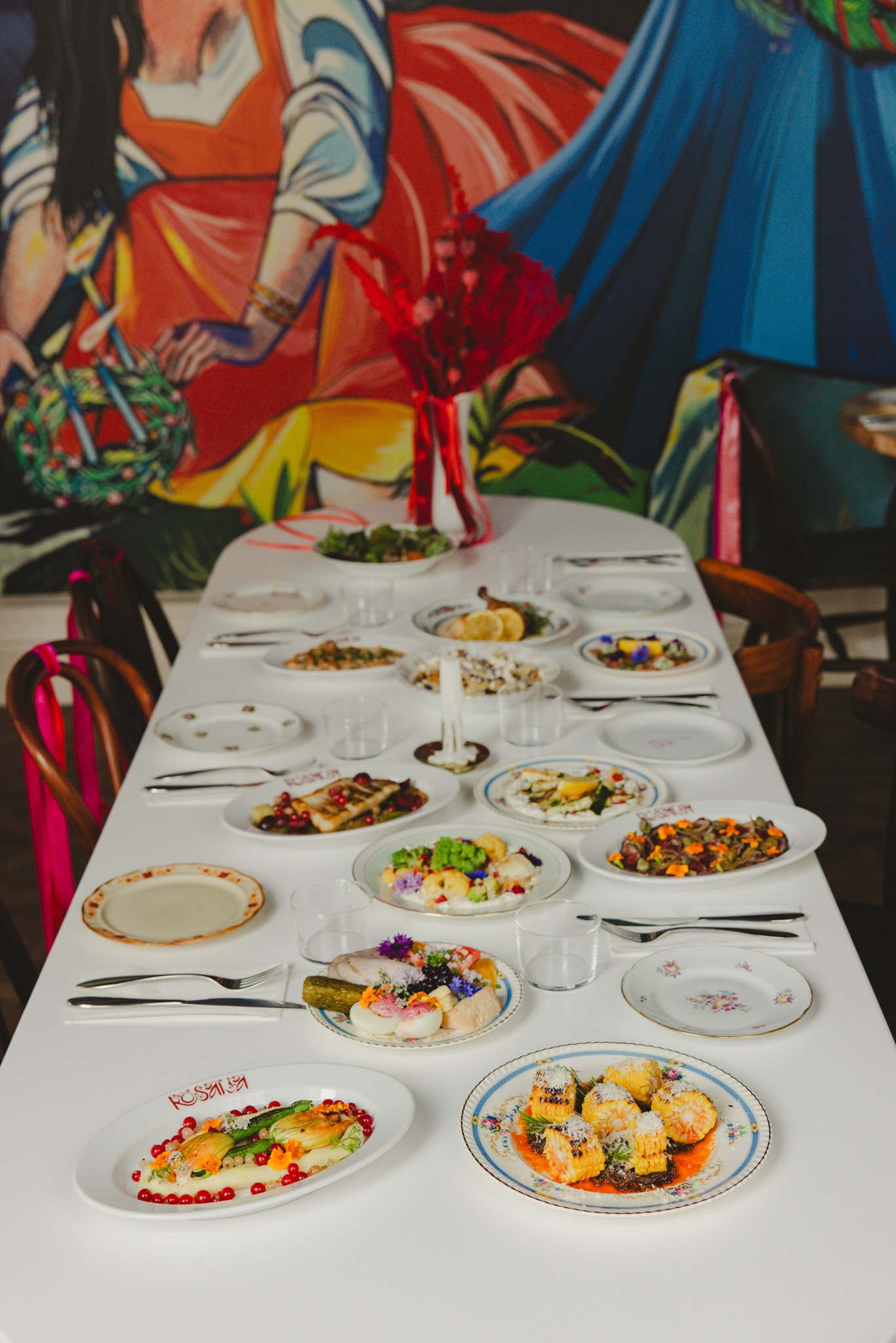 Udekorowany stół z różnorodnymi daniami na talerzach w stylu vintage, kwiaty i czerwone gałązki w wazonie, malowidło na ścianie w tle. Elegancki catering.