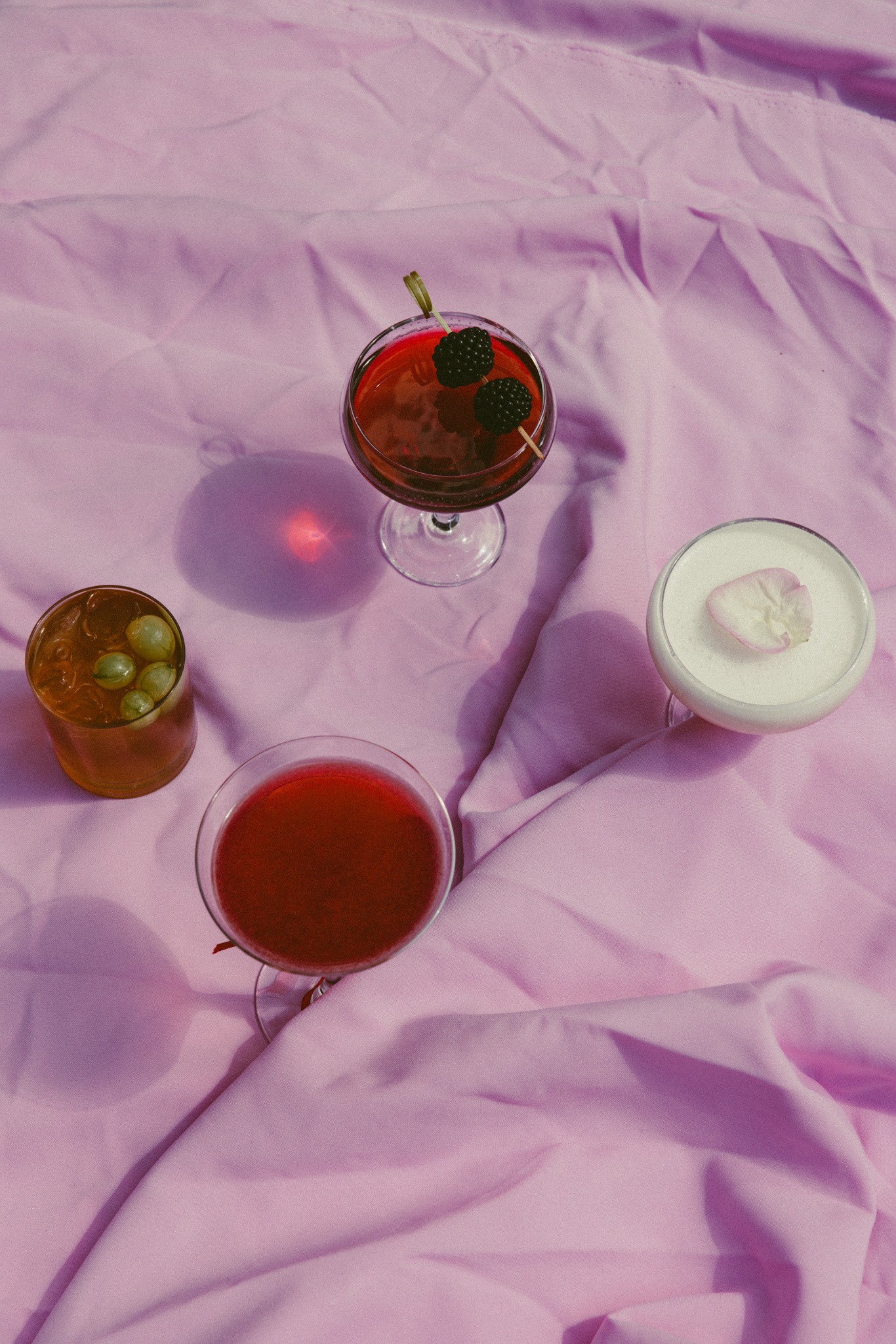 Cztery koktajle o różnych kolorach i dekoracjach na tle pogniecionej, różowej tkaniny. Kreatywna prezentacja drinków z Warszawy.