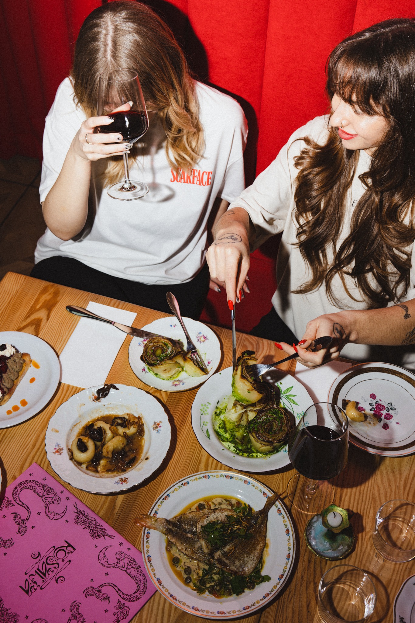 Dwie osoby przy stole z różnorodnymi daniami i winem w restauracji o czerwonym wystroju. Jedna osoba kroi danie, druga trzyma kieliszek wina. Menu leży obok talerzy.