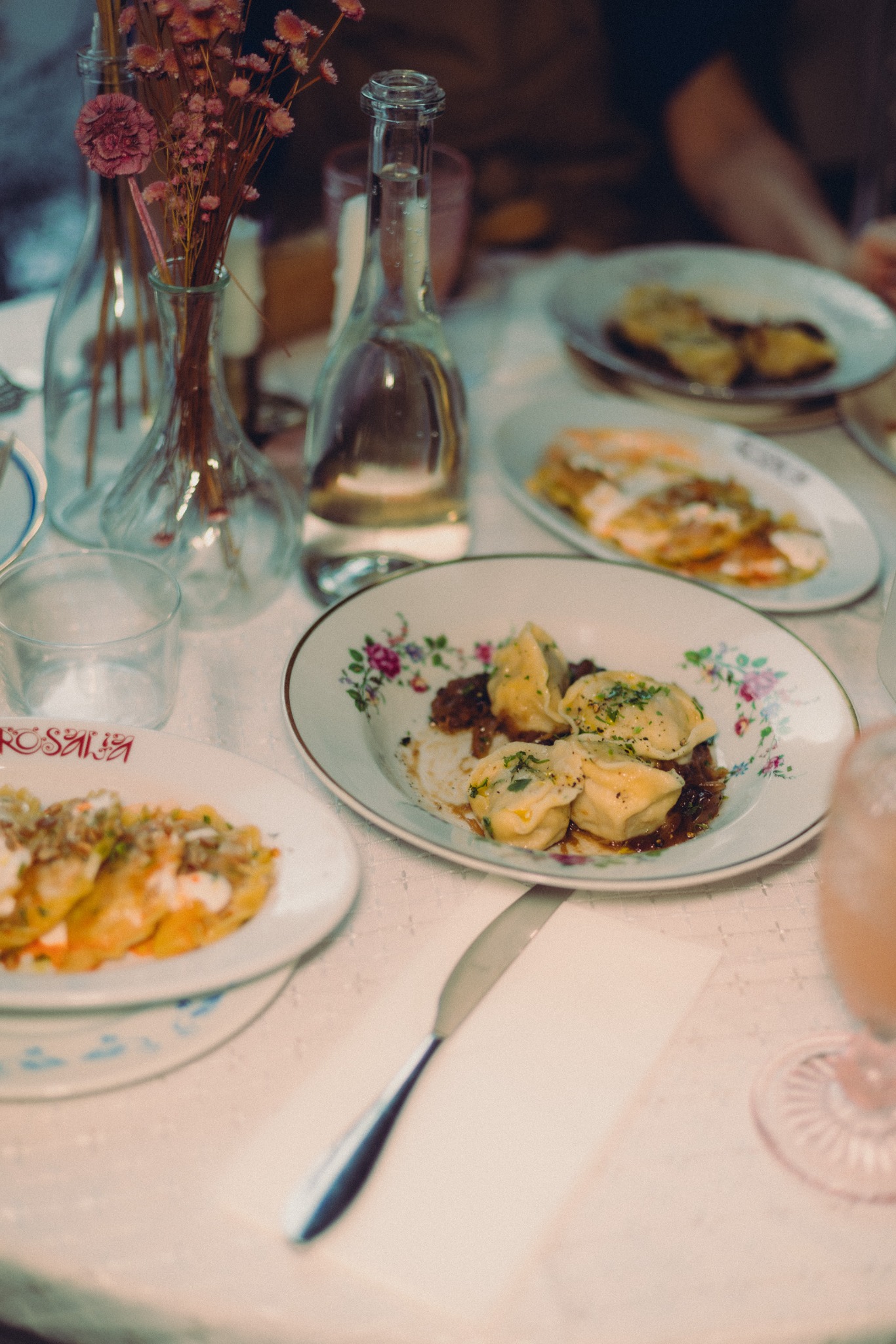 Udekorowany stół z talerzami pełnymi pierogów i placków ziemniaczanych, butelkami wody i wazonami z kwiatami. Romantyczna aranżacja stołu.