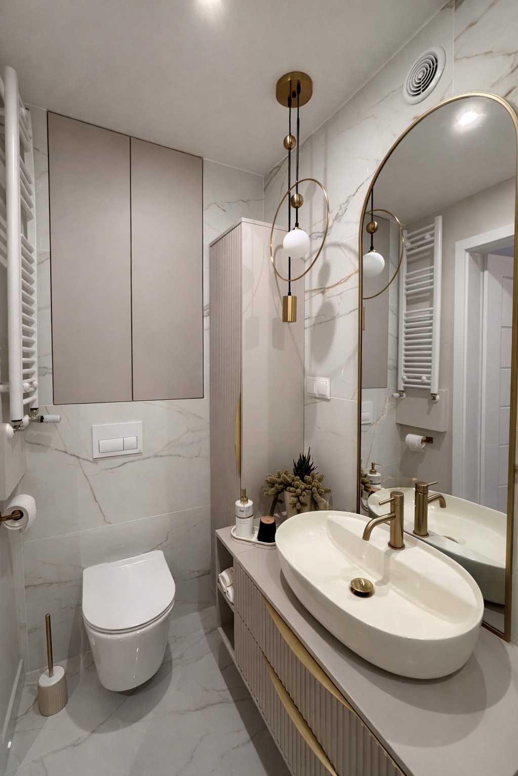 Elegancka łazienka w odcieniach bieli i beżu z marmurowymi płytkami, owalną umywalką nablatową i złotymi akcentami, w tym lustro i lampa wisząca.