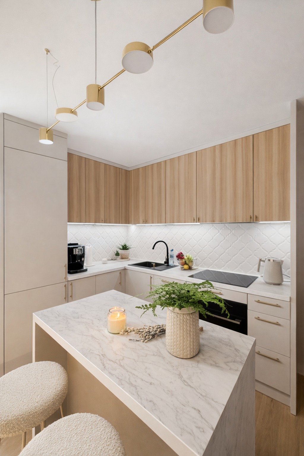 Jasna kuchnia z wyspą i marmurowym blatem. Nad blatem złote lampy. Na wyspie świeca i wazon z paprotką. Ustawione dwa krzesła barowe z jasnym obiciem.