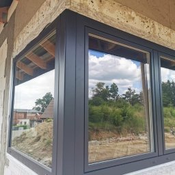 ULTRA ALU DENNIS TUNPRENKAJ - Szare okno narożne z widokiem na zewnątrz, osadzone w surowej ścianie budynku. Widoczne detale montażowe i odbicie krajobrazu w szybach.