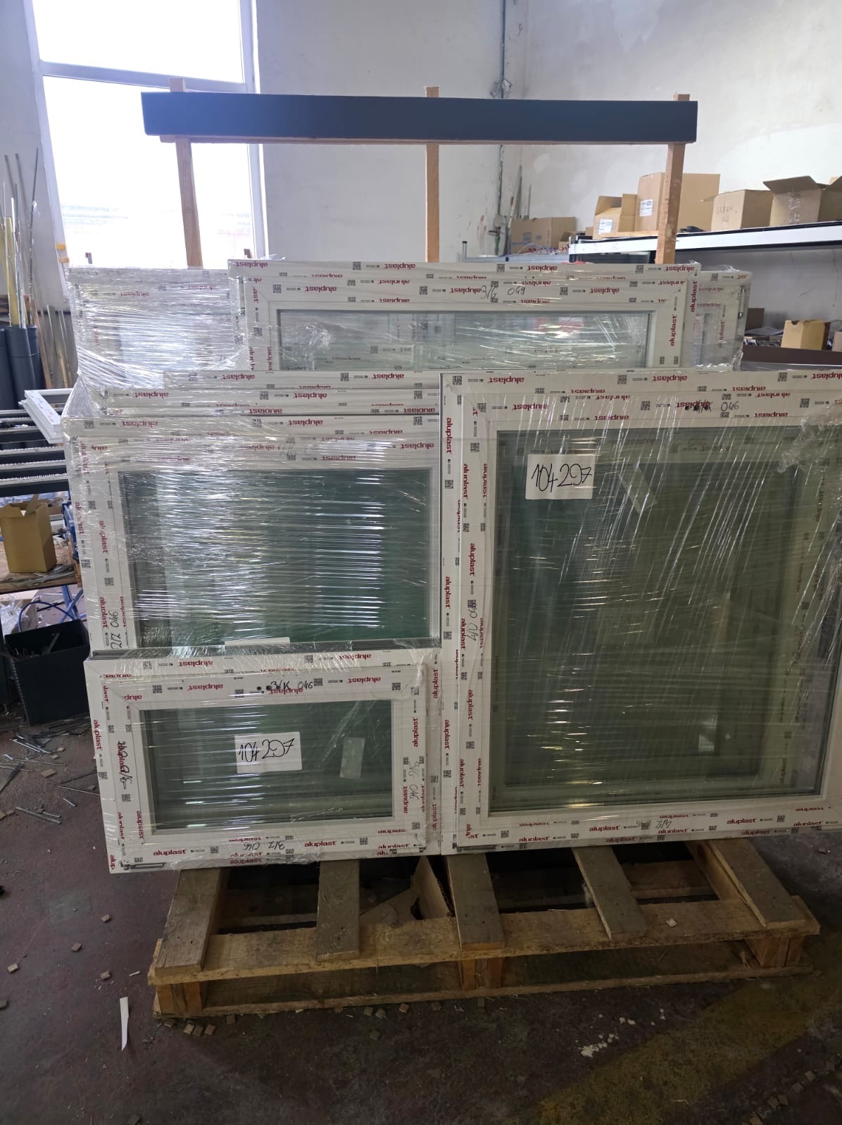 Stos okien PCV Aluplast, owiniętych folią, ułożonych na drewnianej palecie w magazynie. Widoczne etykiety z numerami seryjnymi i oznaczeniami producenta.